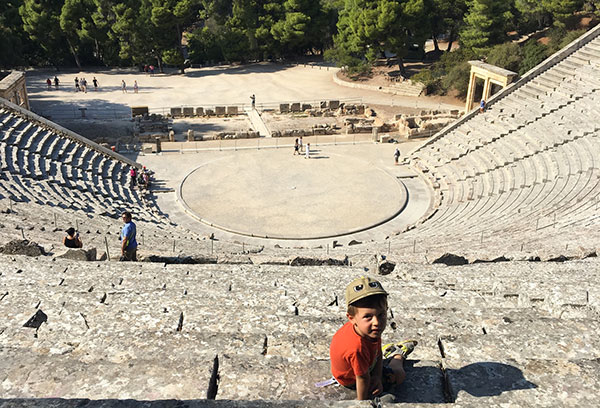 bambino seduto con cappellino siullo sfondo teatro greco antico