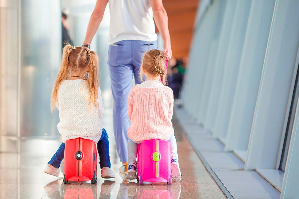 Mamma di spalle trasporta due bambine sulle valigie cavalcabili